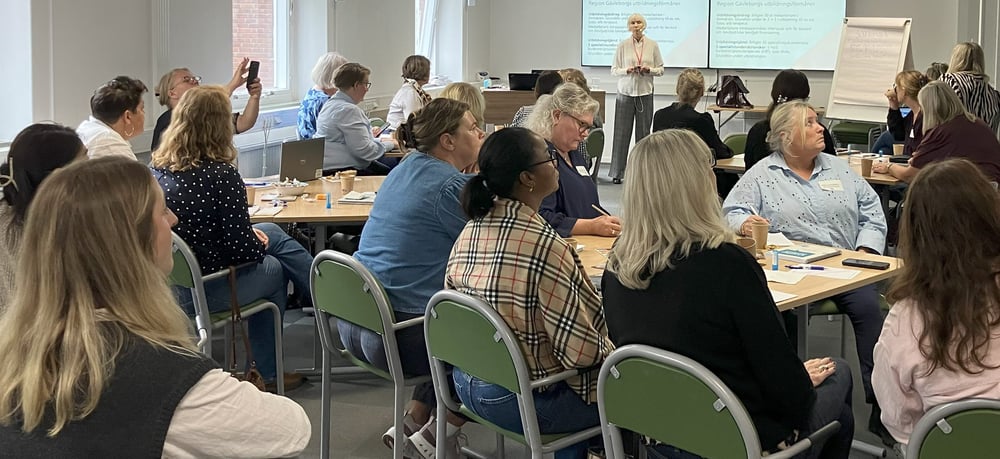 Deltagare sitter vid bord i ett konferensrum och lyssnar på en presentation som visas på storbildsskärm.