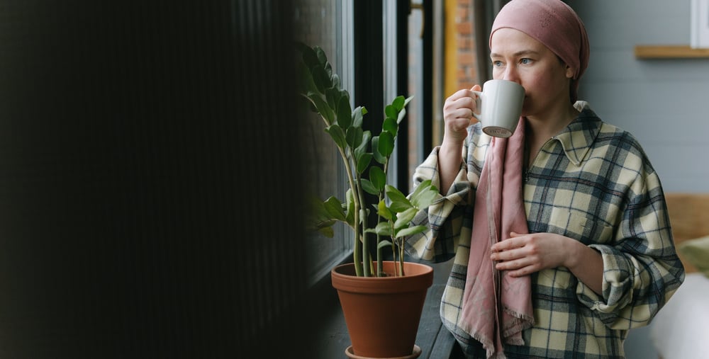Person står vid ett fönster och dricker ur en kopp. I fönstret står en krukväxt.