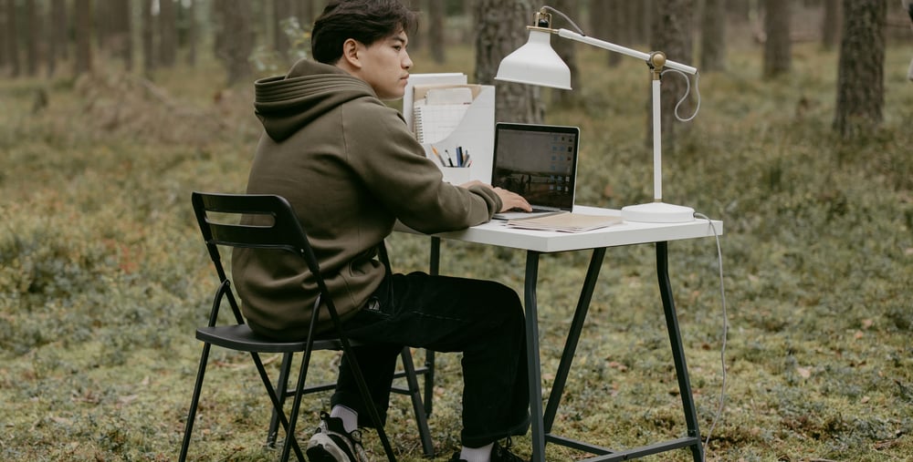 En person sitter vid ett skrivbord i skogen med dator och skrivbordslampa.