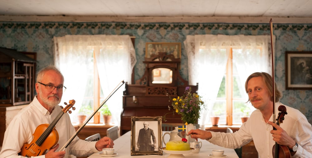 Två personer sitter vid ett bord med varsitt instrument och tittar in i kameran.