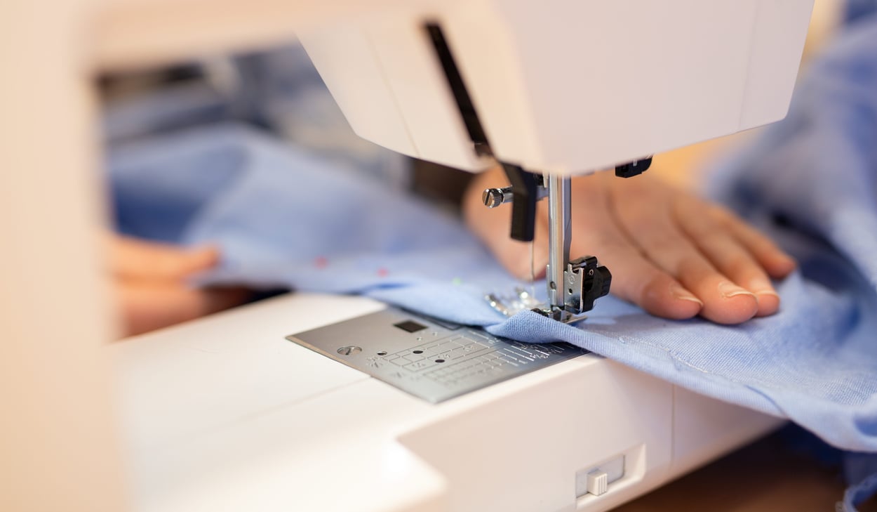 Närbild på en symaskin där en hand styr ett blått tyg genom nålen under sömnad.