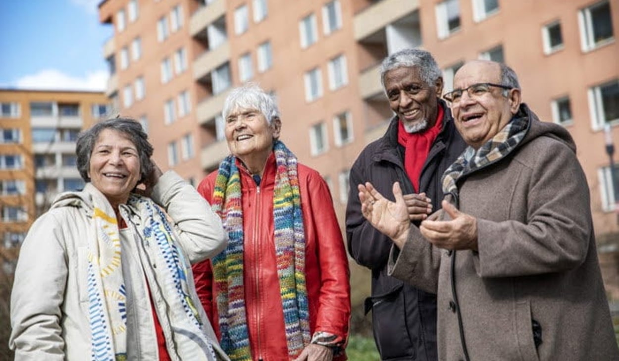 Fyra äldre personer står utomhus och samtalar framför ett bostadshus.