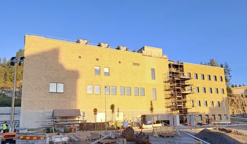 Bilden visar den nya teknikbyggnaden med administrativ byggnad på Hudiksvalls sjukhus. Himlen är blå och molnfri, i förgrunden finns byggarbetsmaterial. Byggnaden är en del av Framtidsbyggets projekt på Hudiksvalls sjukhus. 