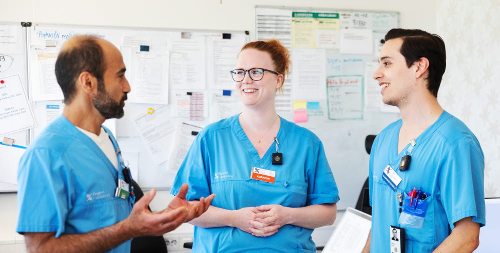 Tre personer i vårdkläder samtalar i ett arbetsrum med whiteboard och datorer i bakgrunden.