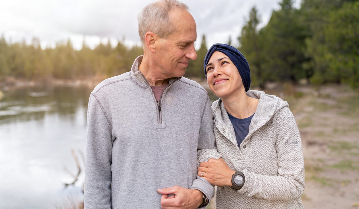 En pappa står och håller i sin vuxna dotter i armkrok. De tittar på varandra varmt och kärleksfullt. De står ute i naturen i en skog och en sjö skymtar i bakgrunden. 