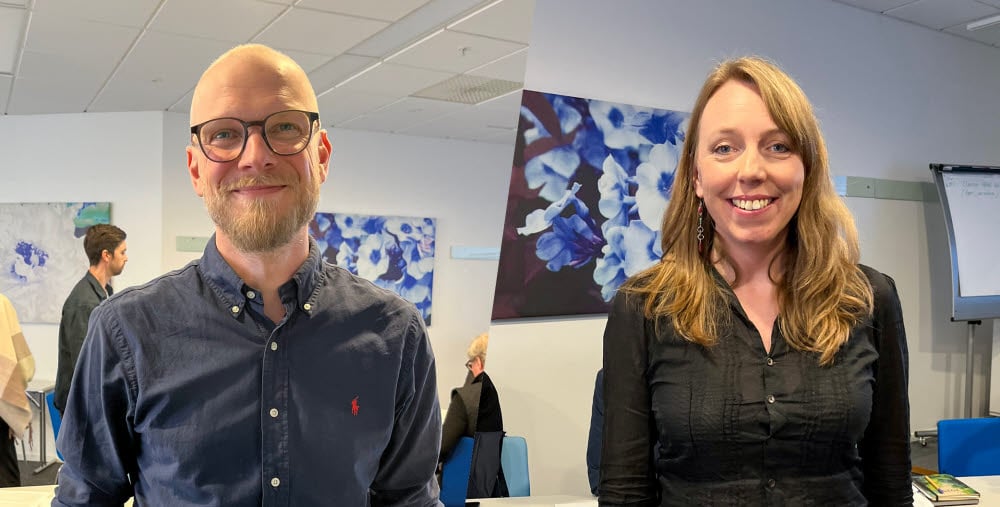 En man med glasögon, skägg och mörkblå skjorta står leende i förgrunden i ett konferensrum. Bredvid står en leende kvinna och i bakgrunden syns en målning med blå och vita blommor samt en blädderblockstavla.