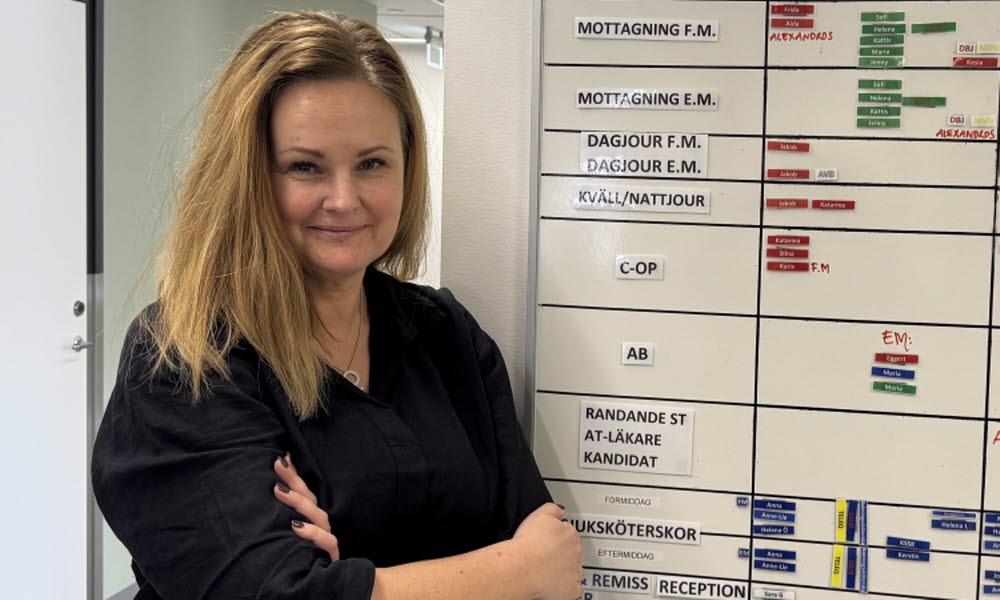 Kvinna står med armarna i kors och lutar sig mot en vägg med en whiteboard med olikfärgade lappar på. Hon tittar in i kameran och ler.