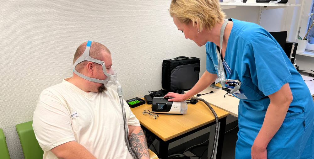 Sjuksköterskan Eva Berglund står och visar en CPAP-apparat åt patienten Rasmus Karlström som sitter ned på en stol. 