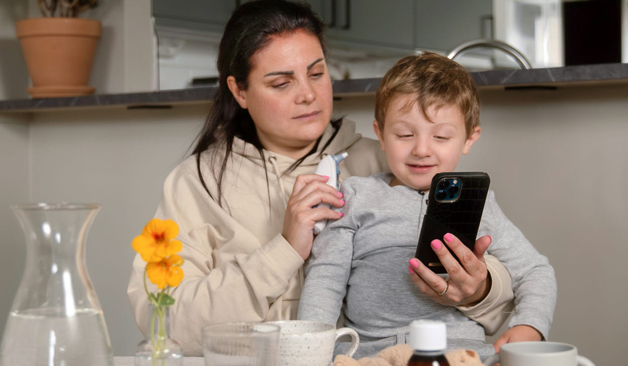 Mamma med barn i knä håller i telefon och termometer