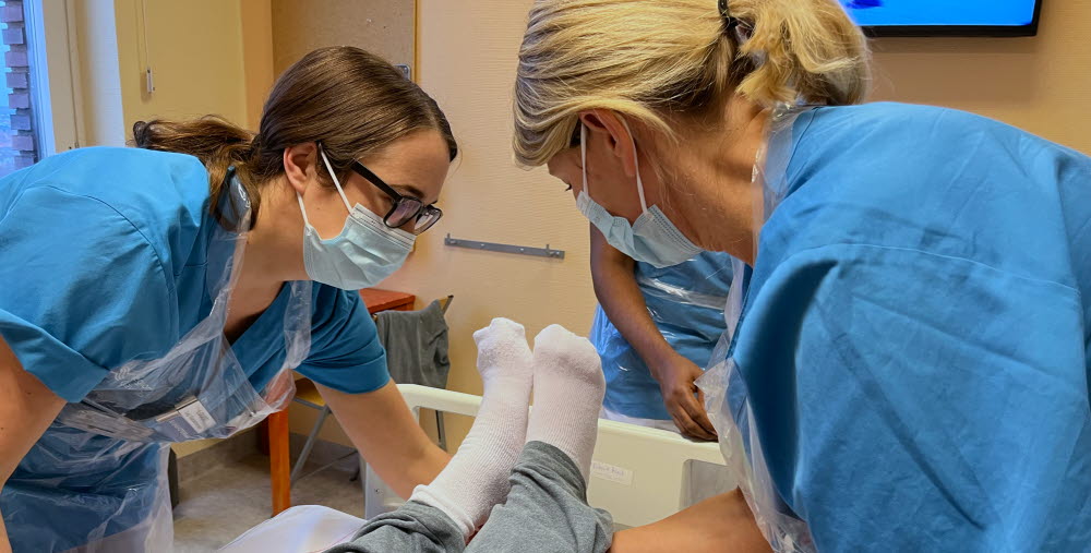 Två vårdpersonal med munskydd står böjd över fotändan i en säng, där det ligger en patient. De lyfter en fot på en patient, foten har vit socka på sig. Endast underbenen syns. 