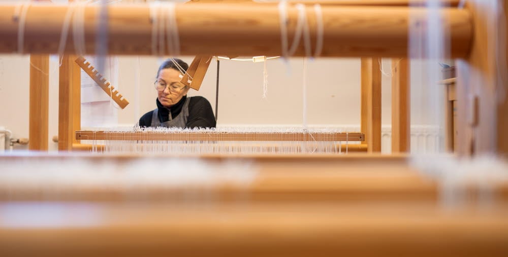 En kvinna syns genom delar av en vävstol medan hon arbetar koncentrerat med vävning.