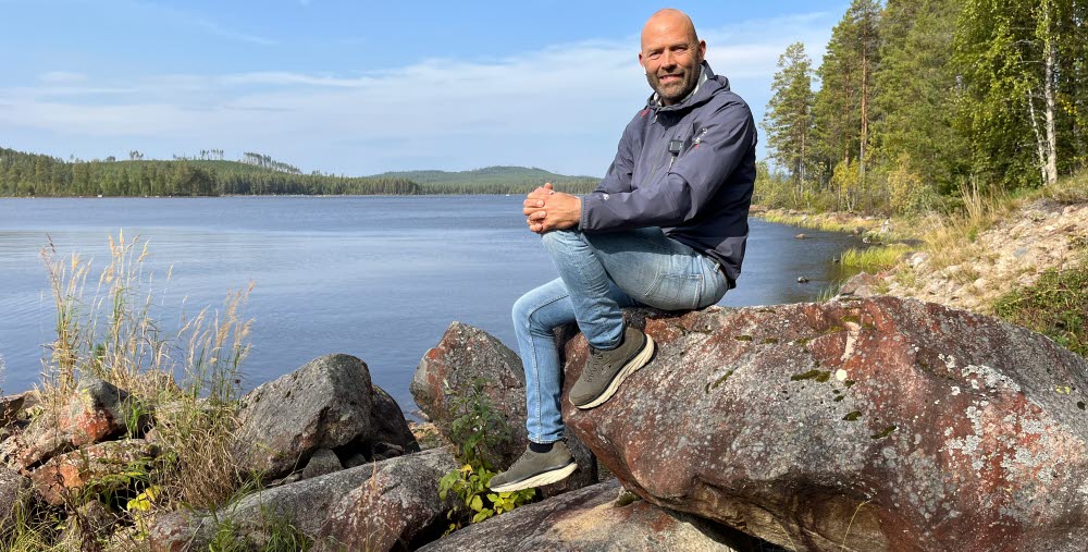 En person i helfigur sitter på sten framför en blå sjö. Sommar.