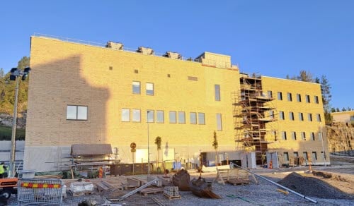 Bilden visar den nya teknikbyggnaden med administrativ byggnad på Hudiksvalls sjukhus. Himlen är blå och molnfri, i förgrunden finns byggarbetsmaterial. Byggnaden är en del av Framtidsbyggets projekt på Hudiksvalls sjukhus.