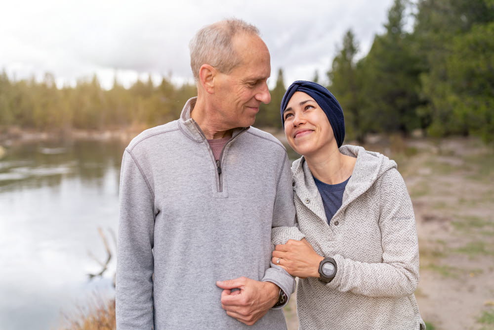 En pappa står och håller i sin vuxna dotter i armkrok. De tittar på varandra varmt och kärleksfullt. De står ute i naturen i en skog och en sjö skymtar i bakgrunden.