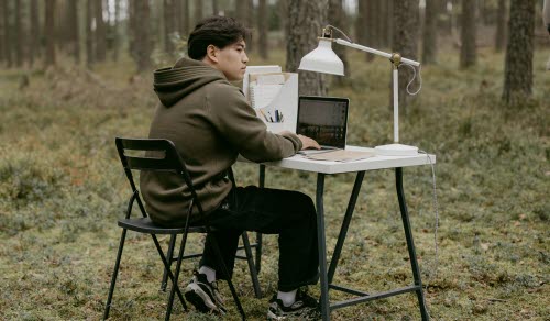 En person sitter vid ett skrivbord i skogen med dator och skrivbordslampa.