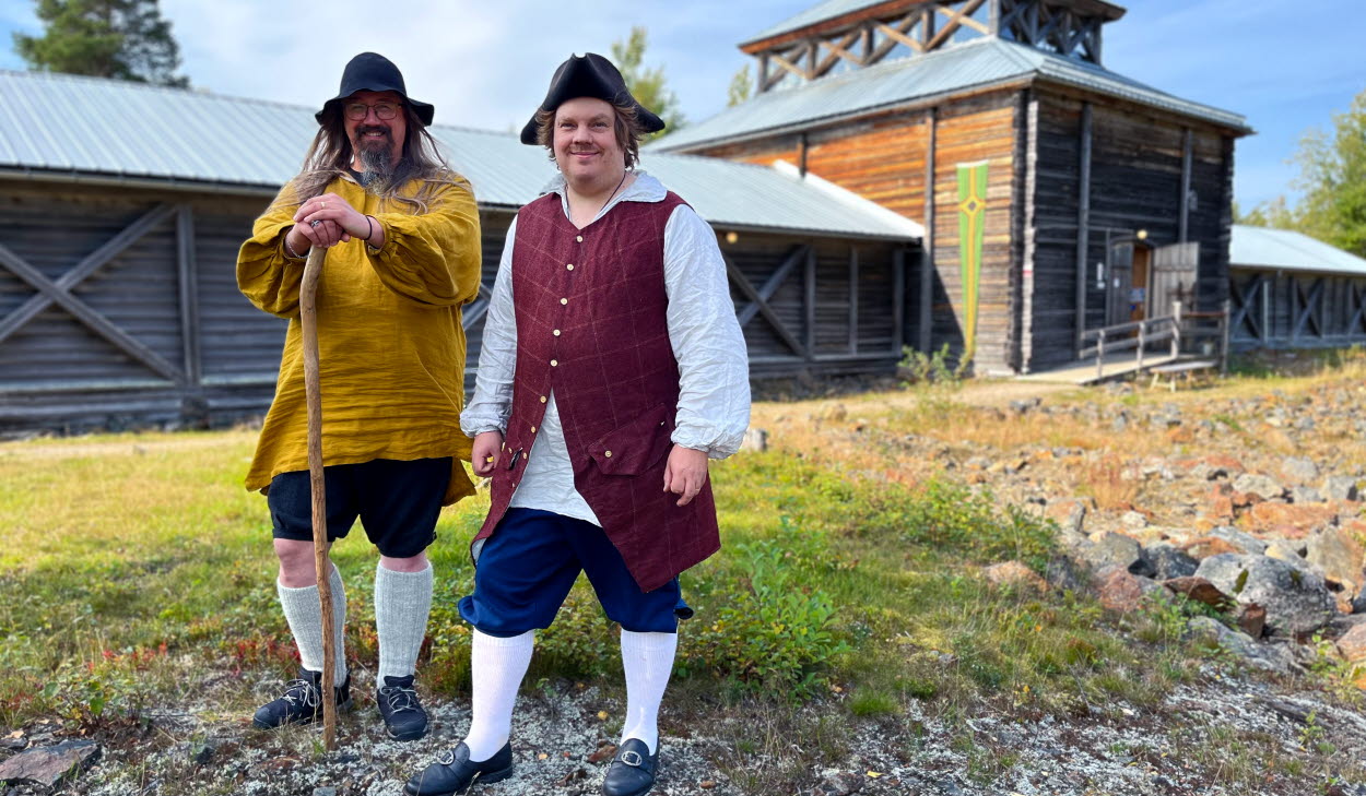 Två män i helfigur klädda i 1700-talskläder står på stenig mark framför en träbyggnad i solsken.
